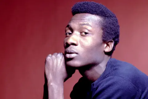 Getty Images Jimmy Cliff is wearing a dark short-sleeved shirt and is posed against a solid reddish-brown background. One arm is bent at the elbow, with the hand resting near the chin, creating a thoughtful posture. The lighting is soft, highlighting the contours of the arm and the texture of the shirt.
