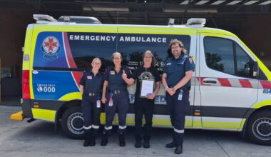 Three paramedics in uniform and one man holding a framed certificate standing in front of an ambulance vehicle.