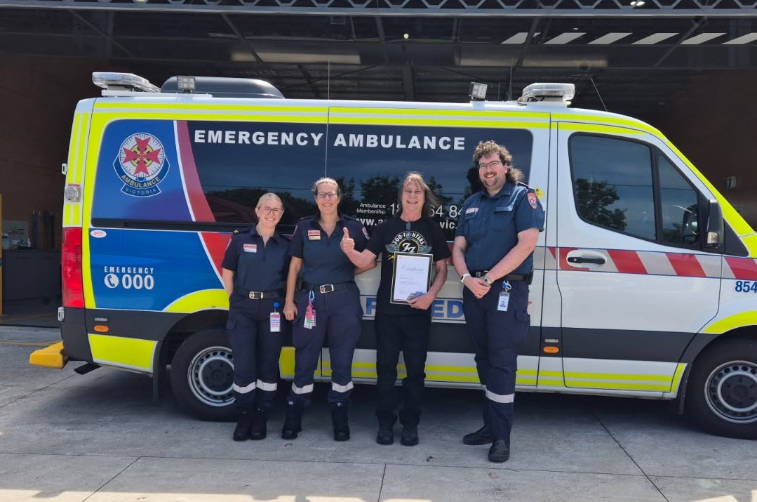 Three paramedics in uniform and one man holding a framed certificate standing in front of an ambulance vehicle.