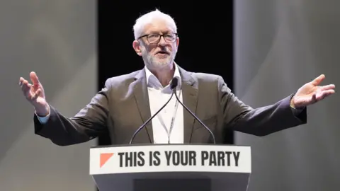PA Media Jeremy Corbyn stands behind a podium on a stage with arms outstretched. The podium has a sign reading ‘THIS IS YOUR PARTY’ in bold black letters with a red triangular graphic on the left. Two microphones are mounted on the podium, and the background is dark with soft lighting.