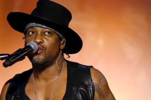 Getty Images D’Angelo is performing on stage, wearing a wide-brimmed black hat and a black leather vest over a deep V-neck top. A silver chain necklace is visible around the neck, and the left arm shows a detailed tattoo design. He is positioned near a microphone with a black foam cover. The background features a warm orange gradient, creating a dramatic and vibrant concert atmosphere.