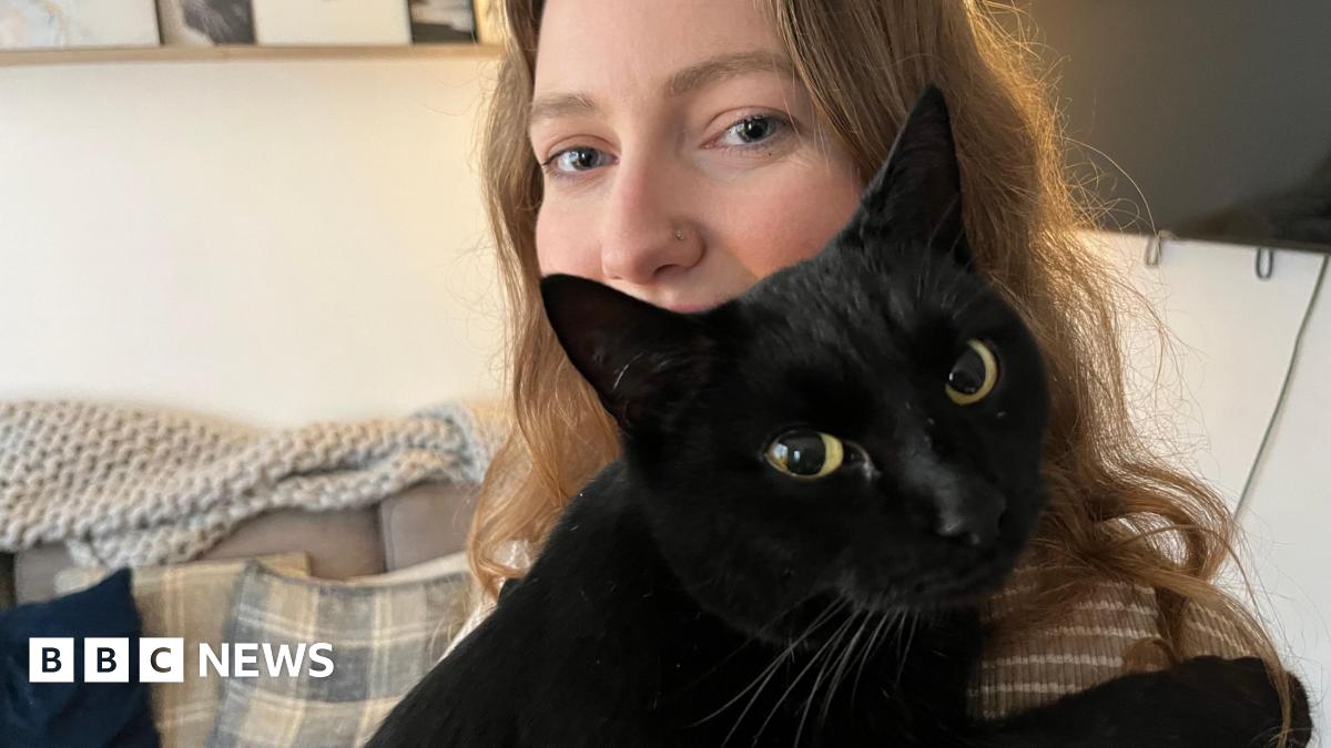 A woman with long brown hair holding her black cat looking at the camera