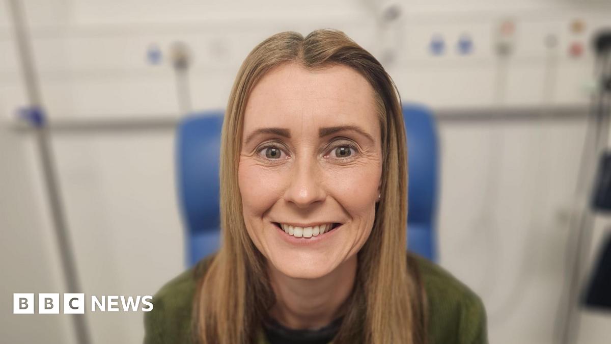 Natalie is smiling at the camera, she has blonde shoulder length hair and is wearing a green jumper. She is sitting in a blue hospital chair.