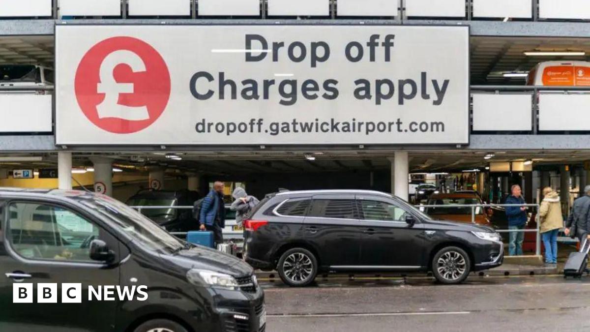 A large sign at an airport car park saying Drop off charges apply with cars and people and cases below it
