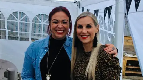 Claire Berridge Lois Sherret smiling to camera, wearing a black top, blue shirt and putting her arm around Claire Berridge who has long, blonde hair and is wearing a leopard print top 