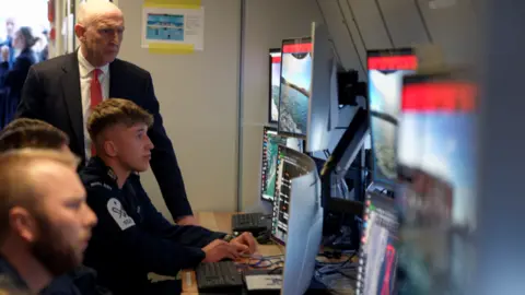 The defence secretary, John Healey looks at the computer screen of a young man in the navy. He is one of three men sitting a desk monitoring several screens. 