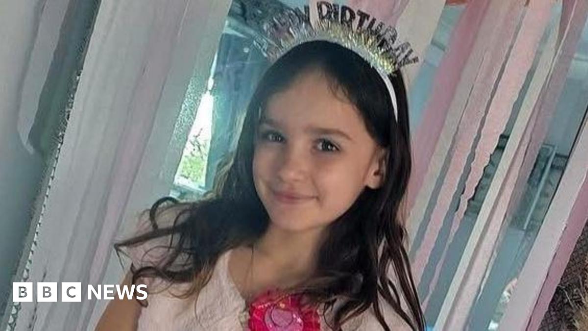 Aria smiles at the camera in front of a doorway filled with pink streamers. She has long brown wavy hair and is wearing a pink dress and shrug. She has a bright pink birthday badge on, and a sparkly headband which reads "HAPPY BIRTHDAY".