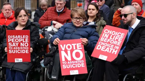 EPA Baroness Tanni Grey-Thompson holds a red placard reading: "Vote no to assisted suicide". She is in a wheelchair and wearing glasses with a blue coat. Two other campaigners are alongside her, holding placards reading "Disabled people say no to assisted suicide" and "Choice in life before choice in death". 