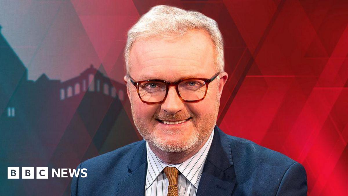 A head and shoulders publicity shot of Peter Henley sitting in a BBC studio. He is wearing a blue suit, white shirt with blue pinstripes and a mustard coloured tie. He has short white hair and a short beard, brown rimmed glasses. The backdrop is predominantly red.