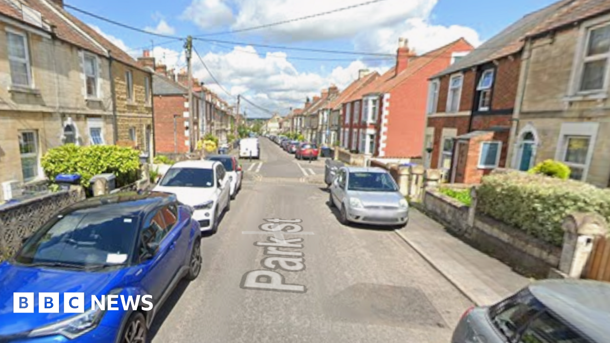 View of a suburban street with cars parked either side of a narrow roadway and clouds in the background.