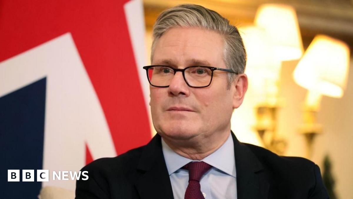 Headshot of Sir Keir Starmer in a suit sitting in front of two bright lamps and a UK flag.