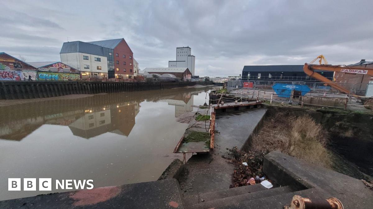 The photo show the rather industrial setting of the River Hull, with concrete steps and discarded rubbish.  An old rusty and broken foot bridge is covered by moss.  The water is dirty and brown.