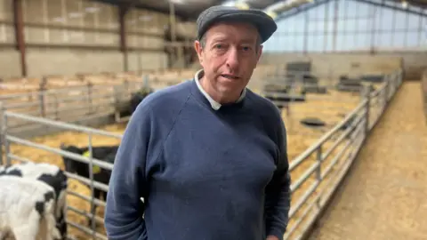 A farmer in a navy jumper is  standing in front of a number of cows in a large shed. He's also wearing a cap.