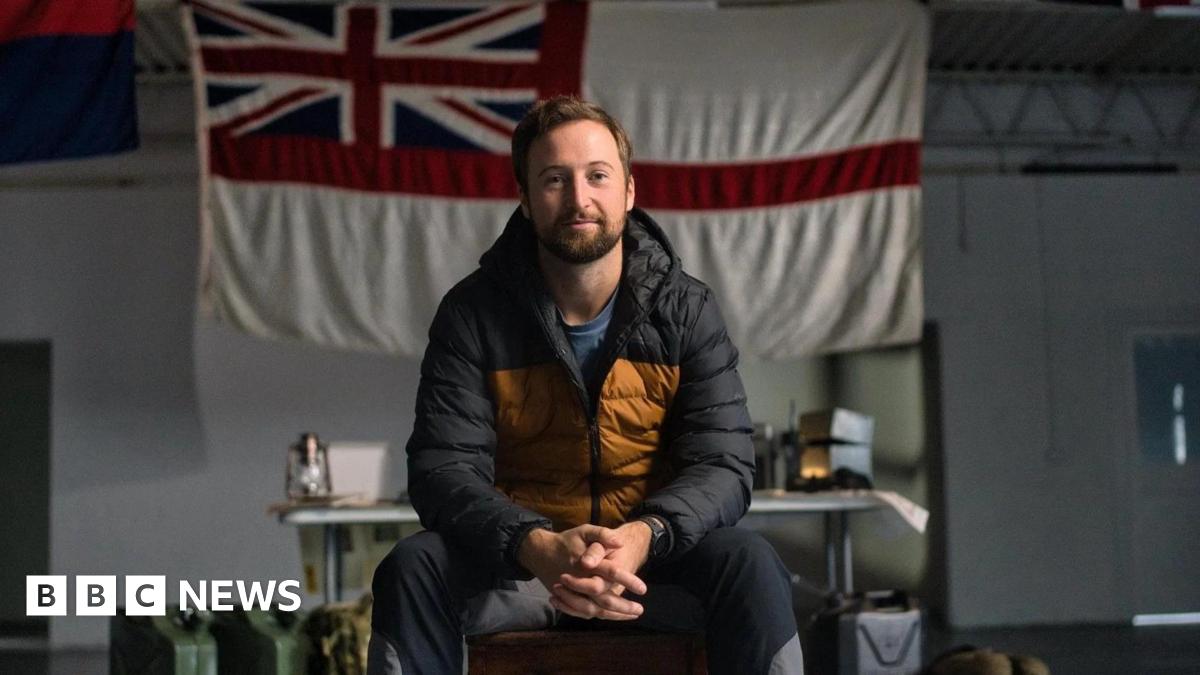 Fred Stokes poses for a photo sitting on a chair, his hands clasped, wearing a black and yellow padded jacket. Behind him is an England flag with the Union Jack in the top left corner. A table with equipment can be seen in the background.