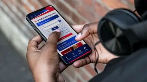 Department for Transport The Great British Railways app being used on someone's phone - it can be seen being held in their hands with the GBR app visible on the screen - an obscured brick work is visible in the near ground 