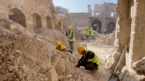 Workers wearing hi-vis vests and yellow helmets clear rubble at the Pasha’s Palace