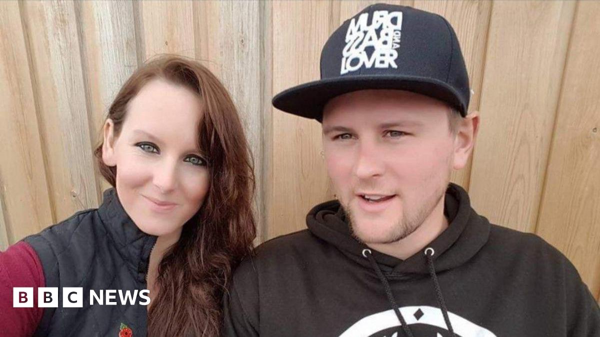 A woman and a man in a selfie. The woman has long brown hair and is wearing a red long-sleeved top under a black gilet. The man is wearing a black hoodie with a large white logo on it, and a black cap with white writing on it