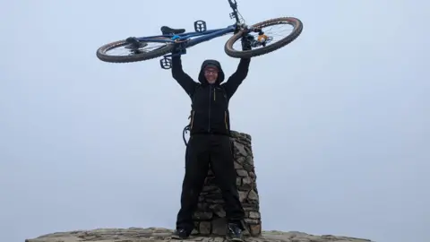 Phil James A man in a black hooded coat and black trousers is standing at the summit of mount snowdon holding a bike above his head
