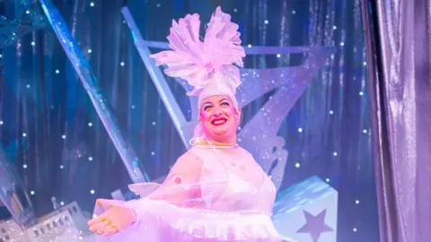 Tron Theatre Johnny McKnight onstage, dressed in a bright pink and white fairy costume. He is grinning and posing.