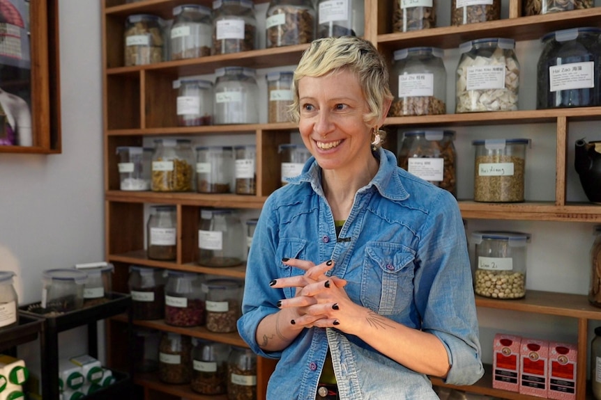 A smiling, short-haired blonde woman stands in a room of shelves packed with labelled jars.