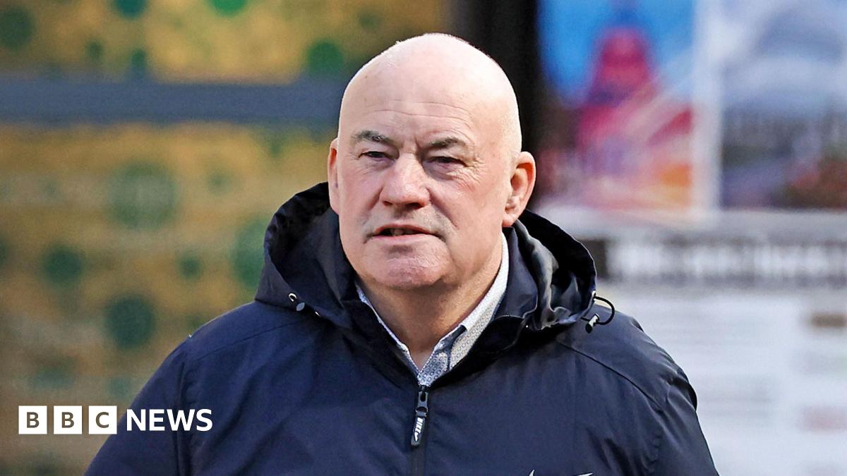 A bald man wearing a black zip-up rain coat over a pale shirt. The background is out of focus.
