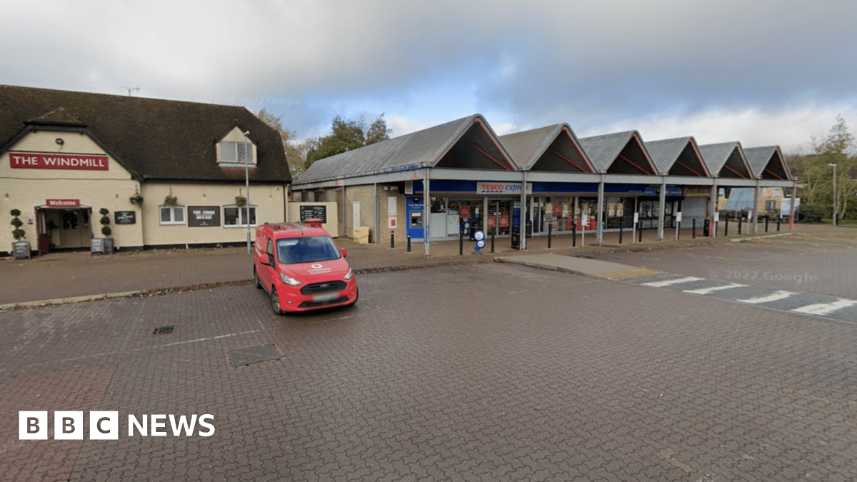 The Freshbrook village centre car park. There is a pub, named the Windmill, to the left hand side, with a neighbouring Tesco express shop. A lone van is parked in the car park.