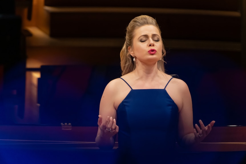 Siobhan Stagg sings on stage with her eyes closed and her hair in a formal up-do. She wears a blue evening gown.