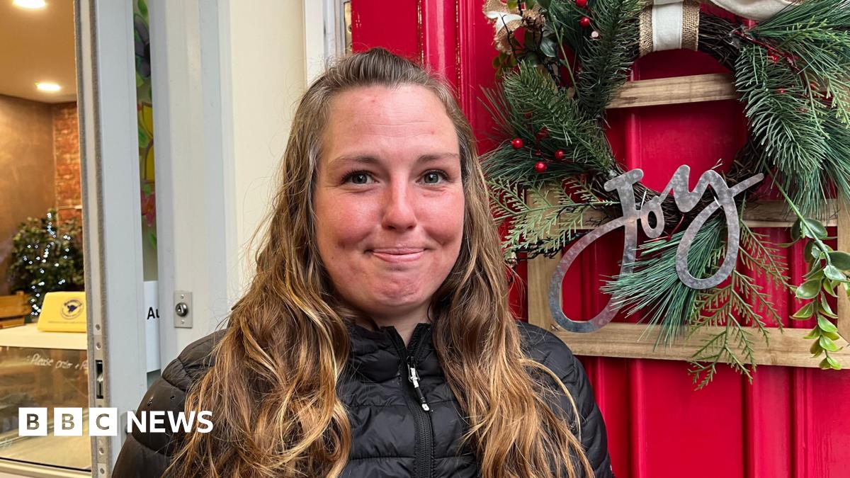 A young woman looking into the camera and smiling slightly. She has long, wavy brown hair and is wearing a black puffer jacket. She's stood in front of a red door that has a green wreath on it, with the word 'Joy' written in silver letters.