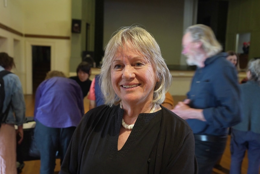 A retirement-aged blonde woman in black blouse and white necklace smiles as people move around behind her.