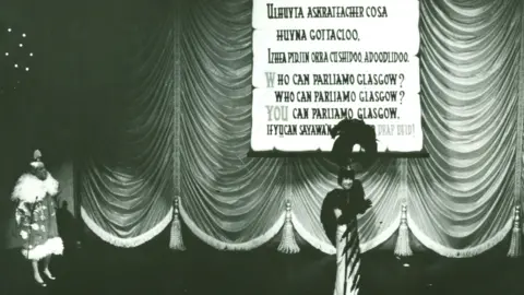 King's Theatre A pantomime production. Two people are onstage dressed in elaborate costumes, while a song sheet has been dropped down behind them, with words for the crowd to join in with