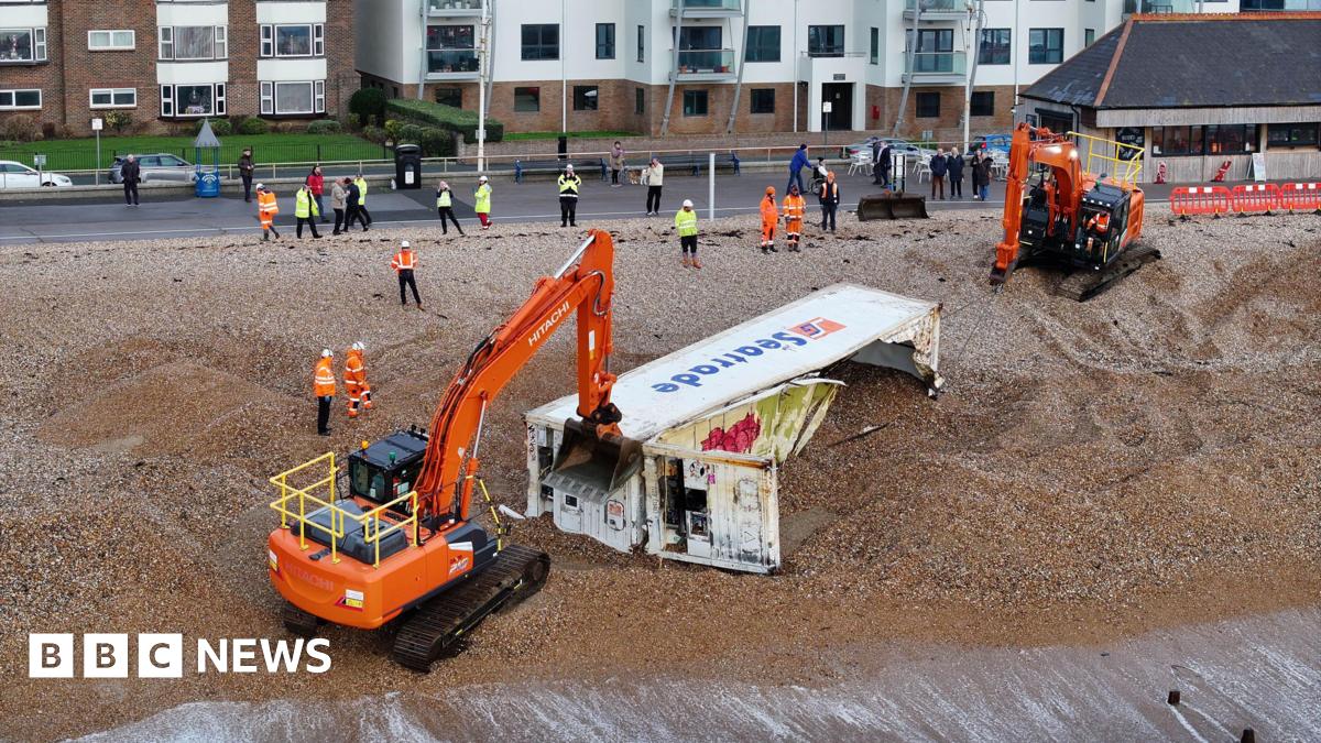 Two orange excavators on the beach removing shingle from a white shipping container so they can move it