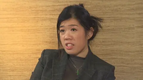 Claire Lai with her hair in a bun, wearing a grey blazer. She is in the middle of speaking.
