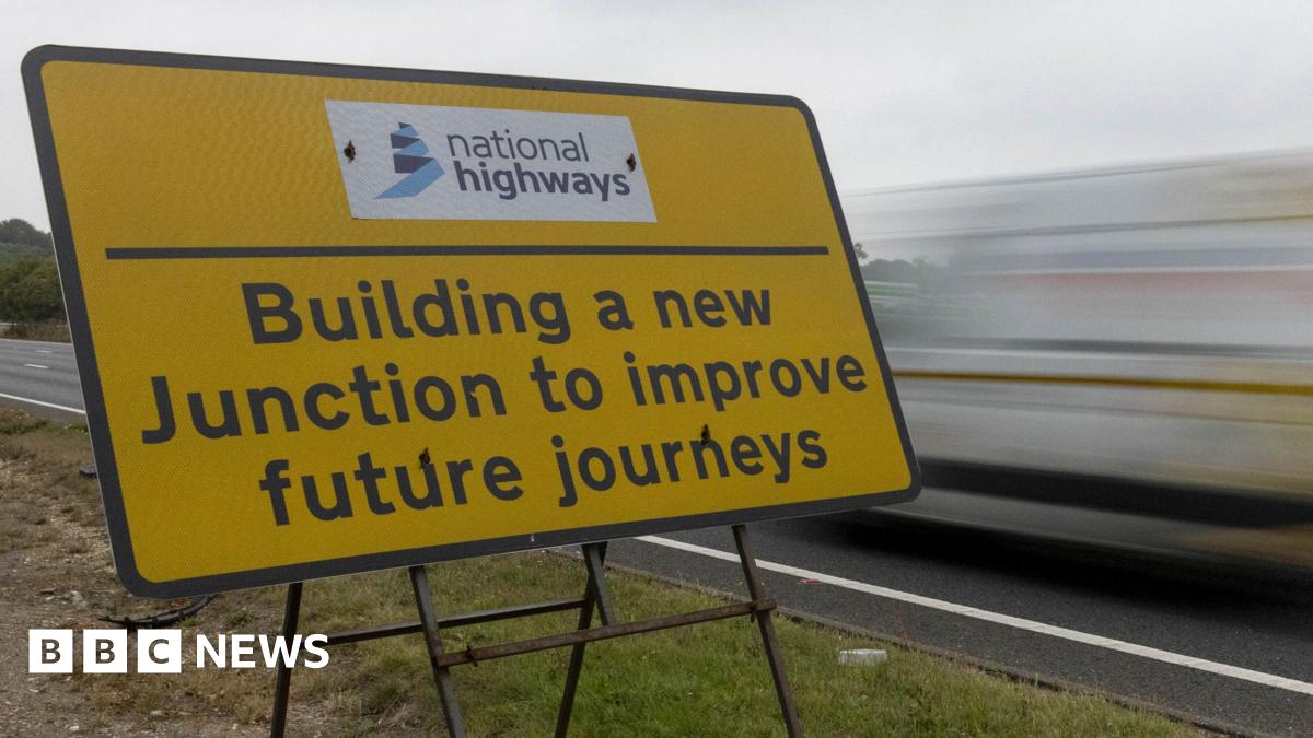 Close up of a yellow sign on a grass verge with the words BUILDING A NEW JUNCTION TO IMPROVE FUTURE JOURNEY written in black. A blurred vehicle drives past the sign on the right