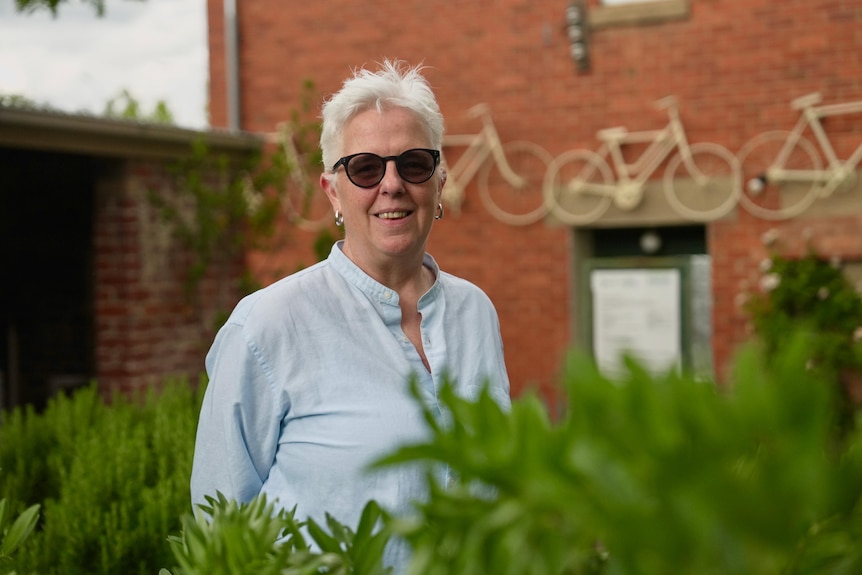 A short-haired woman in sunglasses stands in a walled garden and smiles at the camera.  