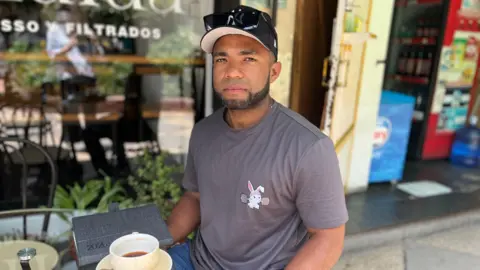 Gabriel, wearing a grey T-shirt with a bunny lifting weights, sits at a cafe table. In front of him are a notebook and a half-drunk cup of coffee. He is wearing a baseball cap on which his sunglasses are perched. 
