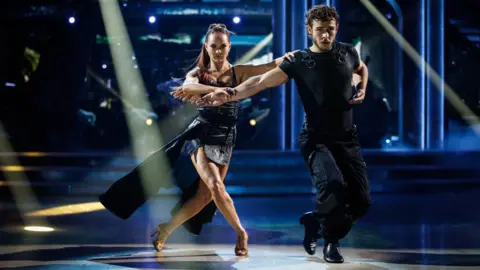 George and Alexis on the dancefloor with their arms out. They are both wearing black and staring straight ahead.