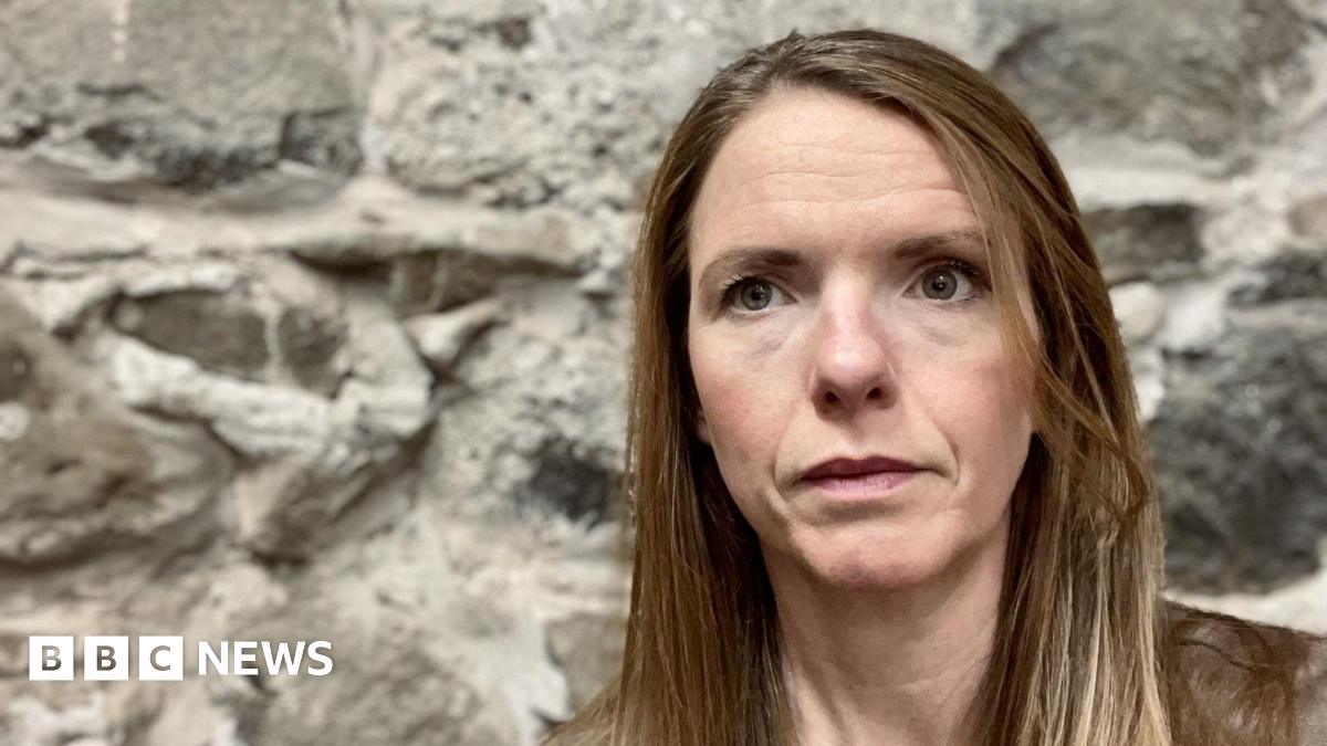 A headshot of Victoria Nichols, who has long brown hair, she is sitting in front of a grey stone wall and is looking slightly off to the left of the camera.