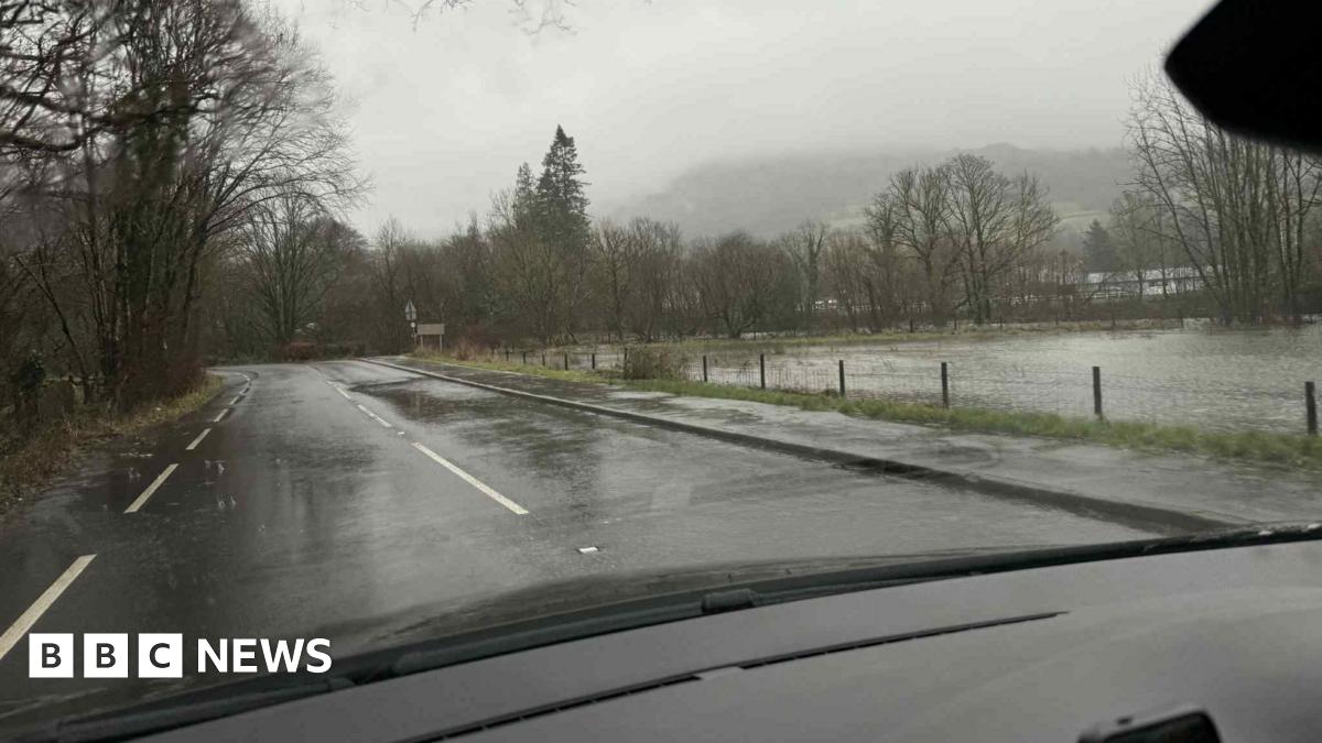 Egremont town councillor David Wright who stands in front of a swollen river under an old bridge. He is wearing a black coat, black hat and grey hoodie. It is raining heavily.