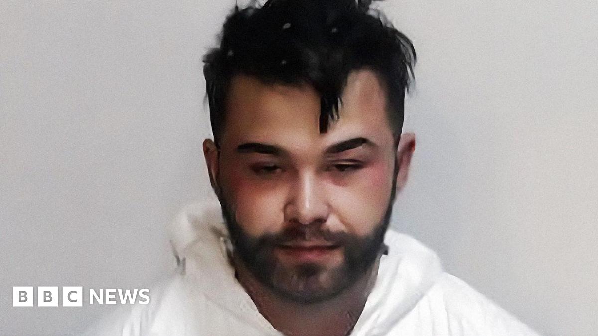 A young man with dark curly brown hair standing by the side of a road. He is wearing a black t-shirt and behind him are yellow crops and blue skies.