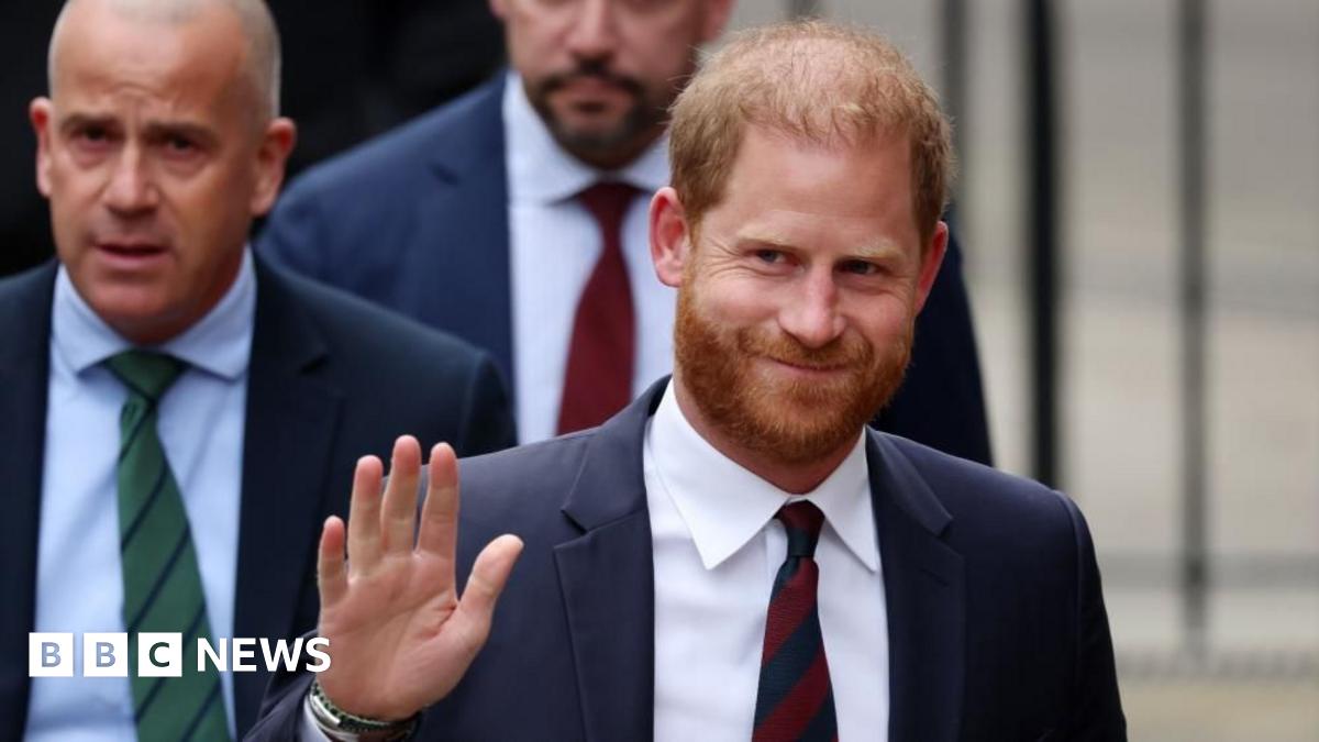 A smiling Meghan Markle stands next to Prince Harry, who wears a blue suit and tie with a white shirt.  She wears long gold-coloured earrings.