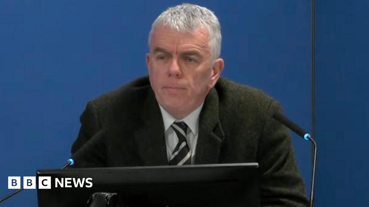 A man with tie and dark jacket sits in front of two microphones and a monitor