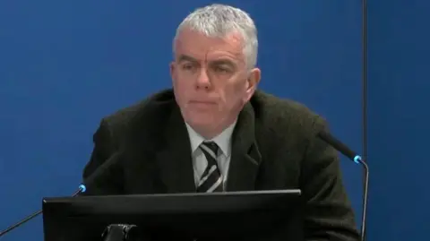 The Undercover Policing Inquiry A man with tie and dark jacket sits in front of two microphones and a monitor 