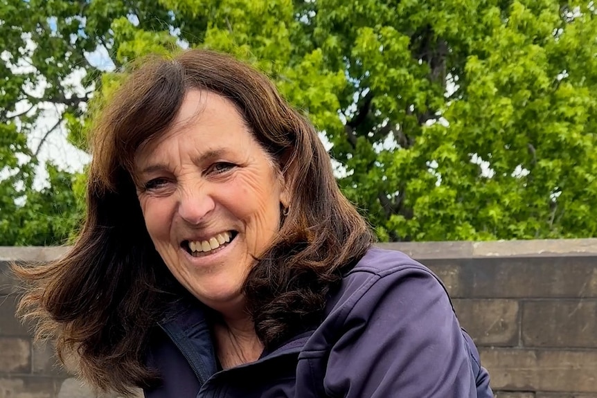Michelle laughs while sitting on a bench there are trees behind her.