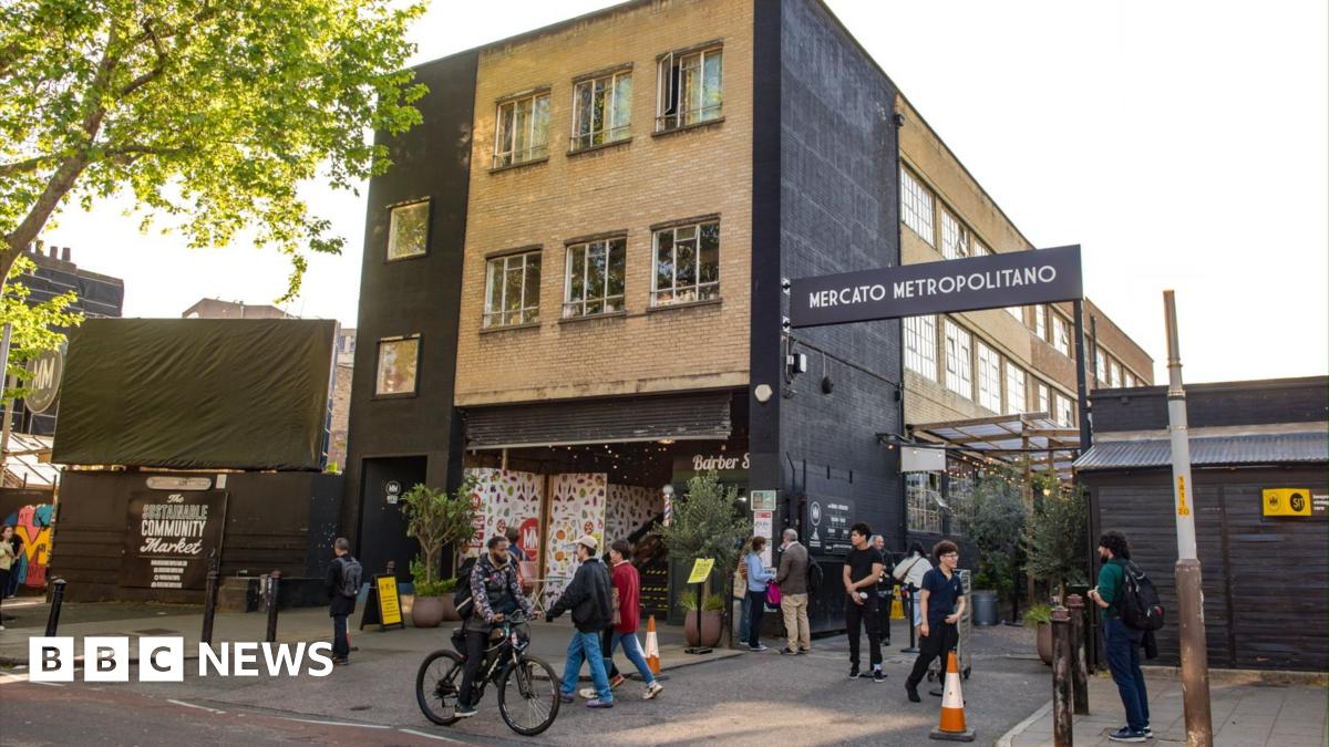 A lively urban street scene featuring Mercato Metropolitano’s entrance, brick building facade, greenery, signage, and people walking and cycling nearby.