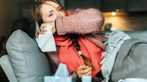 Getty Images An unwell woman lies on under blankets on her sofa and covers her mouth as she coughs. 
