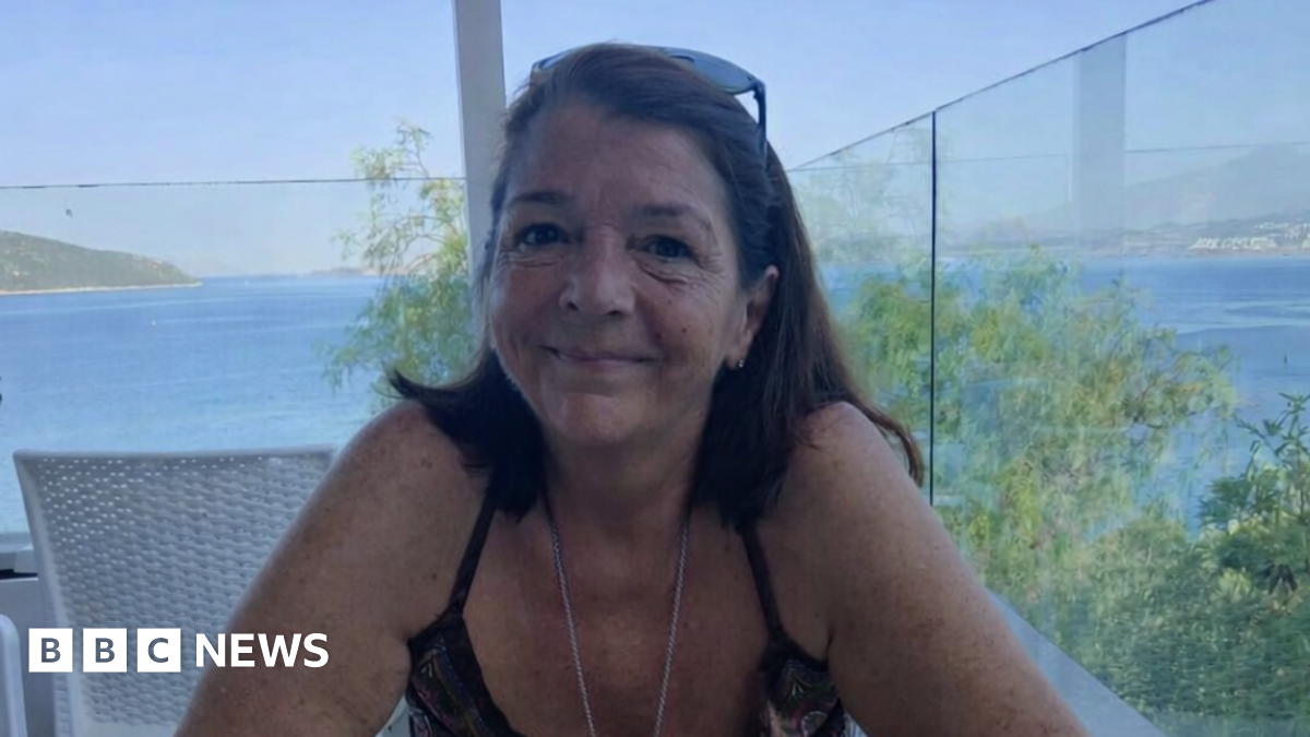 Julie Wilson is pictured with shoulder-length brown hair sitting at a table on a terrace overlooking the sea, smiling at the camera, with blue water and a distant coastline visible behind her.