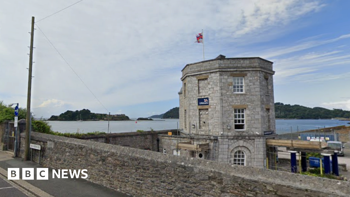 Exterior of a lifeboat station. A small brick wall runs alongside a pavement. There is a stone grey building with multiple windows and lifeboat signage displayed on its outside wall. A calm sea is in the background. Trees are in the distance.