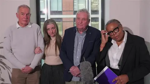 A woman and two men linking arms, standing in front of a window. There is a woman at the end of the row, holding paperwork and standing in front of a microphone. 