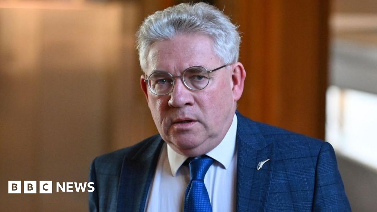 Kenneth Gibson, who has short grey hair and glasses, looks at the camera. He is wearing a blue suit and tie with a white shirt, with wood panelling in the background.