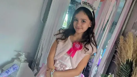 Family photograph Aria Thorpe standing in a white party dress with a pink rosette pinned on it. She is also wearing a silver tiara and has shoulder-length dark brown hair
